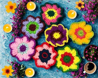 May include: A collection of colorful, handmade crochet flower coasters in various shades of pink, yellow, purple, and red. The coasters are arranged on a blue surface, with candles, flowers, and a cup of coffee.