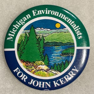 May include: A round button with a green border and the words "Michigan Environmentalists" in white. The center features a landscape with a lake, trees, and mountains. The bottom half is blue with the words "FOR JOHN KERRY" in white.