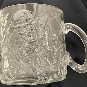 May include: Clear glass mug with a handle. The mug features a detailed embossed image of a character wearing a hat and coat. The design is visible on the front and sides of the mug. The mug is transparent and appears to be made of glass.