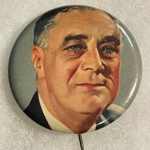 May include: A vintage button featuring a portrait of a man with light blue eyes, likely a political figure. The button has a pin on the back and a glossy finish. The portrait is set against a gray background.