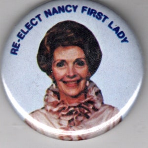 May include: A white button with blue text that reads "Re-Elect Nancy First Lady". A woman with brown hair is pictured in the center of the button.