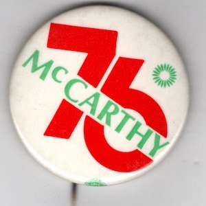 May include: A white button with the text "McCarthy 76" in green and red. The number "76" is in red, and the name "McCarthy" is in green, with a small green floral design. The button has a pin on the back.