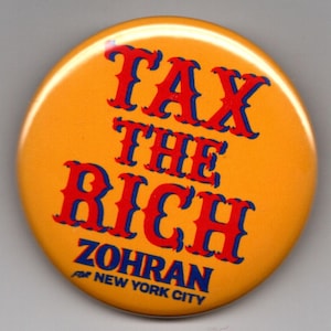 May include: A round, orange button with the text "TAX THE RICH" in red, outlined in blue. Below, it reads "ZOHRAN FOR NEW YORK CITY" in blue. The button is a political statement.