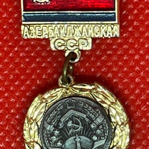 May include: A vintage medal with a red and gold design. The top section features a red background with a gold hammer and sickle emblem. The bottom section is a circular medal with a detailed emblem surrounded by a gold wreath. The text "АЗЕРБАЙДЖАНСКАЯ ССР" is visible.