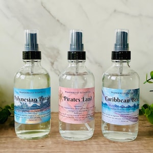 May include: Three clear spray bottles with black spray nozzles and labels. The labels read "Polynesian Paradise", "Pirates Land", and "Caribbean Bead". The bottles are filled with a clear liquid and sit on a wooden surface.