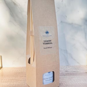 Puede incluir: Una caja de cartón marrón con una ventana transparente que muestra una botella de vidrio de difusor de caña. La caja tiene una etiqueta blanca con el texto "Wanderlust" y "Lemon Verbena Reed Diffuser".