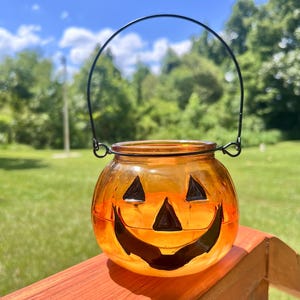 May include: An orange glass jack-o'-lantern jar with a black handle. The jar has a carved face with triangular eyes and a smiling mouth. The jar is sitting on a wooden surface with a green lawn and trees in the background.