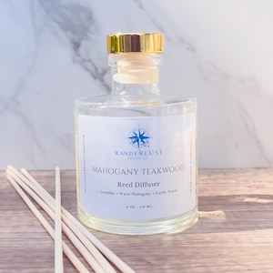 May include: A clear glass bottle with a gold lid and a white label. The label reads "Wanderlust Scents Co. Mahogany Teakwood Reed Diffuser Lavender + Warm Mahogany + Earthy Wood 4 oz / 118 ml". The bottle is sitting on a wooden surface with wooden sticks.