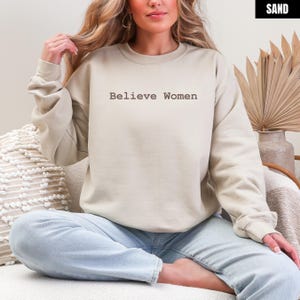 May include: A sand-colored sweatshirt with the text "Believe Women" in a simple font. The long-sleeved crew neck top is worn by a person sitting cross-legged. The background includes a neutral-toned pillow and decorative dried plants.
