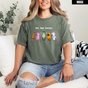 May include: Moss green t-shirt with the text "We Are Antifa" above a row of cartoon characters. The characters include a dinosaur, axolotl, frog, a yellow creature, and a unicorn. The shirt is a casual style.