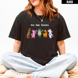 May include: Black t-shirt with the text "We Are Antifa" above a row of cartoon characters including a dinosaur, axolotl, frog, pear, and unicorn. The shirt is a dark color.
