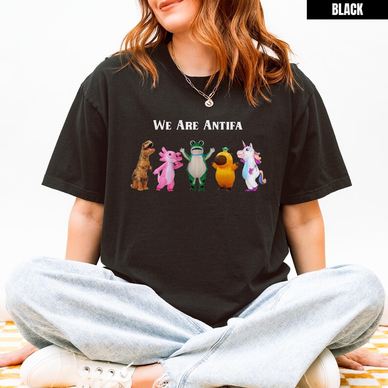 May include: Black t-shirt with the text "We Are Antifa" above a row of cartoon characters including a dinosaur, axolotl, frog, pear, and unicorn. The shirt is a dark color.