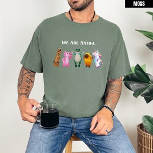 May include: Moss green t-shirt with the text "We Are Antifa" above a row of cartoon characters. The characters include a dinosaur, axolotl, frog, acorn, and unicorn. The shirt is a relaxed fit.