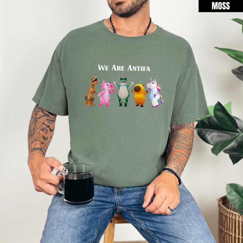 May include: Moss green t-shirt with the text "We Are Antifa" above a row of cartoon characters. The characters include a dinosaur, axolotl, frog, acorn, and unicorn. The shirt is a relaxed fit.