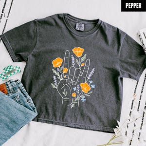 Könnte beinhalten: Dunkelgraues T-Shirt mit Rundhalsausschnitt und einem Handgesten-Motiv mit orangefarbenen und blauen floralen Akzenten. Das Wort "pepper" befindet sich in einem schwarzen Feld oben rechts. Das Shirt wird mit einer blauen Jeans und einer grün-weißen Haarspange kombiniert.