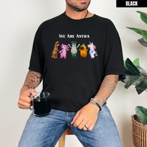 May include: Black t-shirt with the text "We Are Antifa" above a row of cartoon creatures. The creatures include a dinosaur, axolotl, frog, a yellow creature, and a unicorn. The shirt is worn by a person.