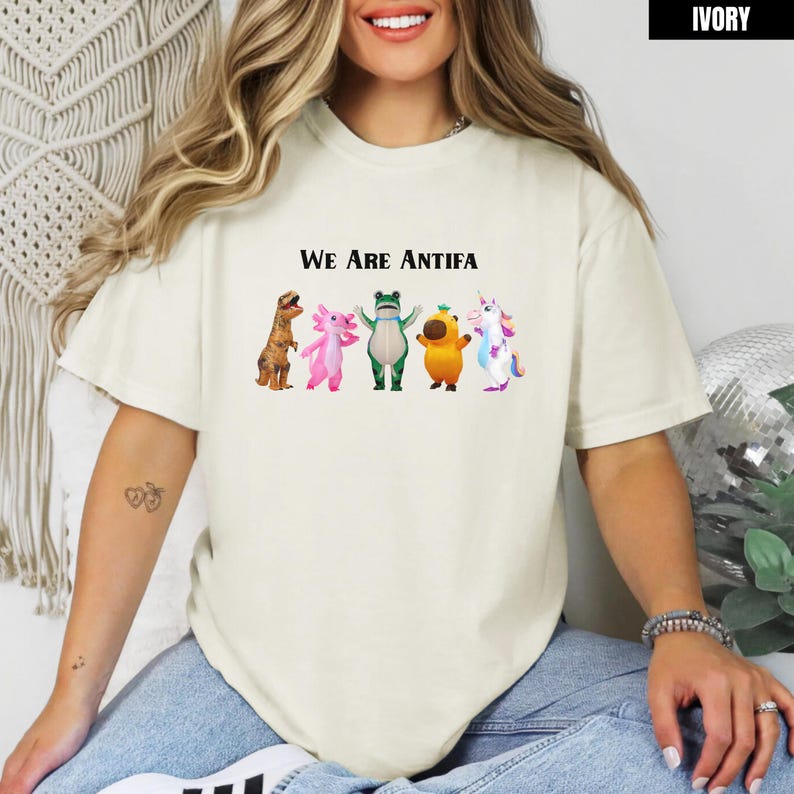 May include: Ivory-colored t-shirt with the text "We Are Antifa" above a row of cartoon characters: a dinosaur, axolotl, frog, mushroom, and unicorn. The shirt is a relaxed fit.