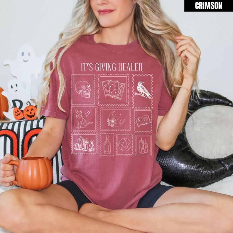 May include: Crimson-colored t-shirt with the text "IT'S GIVING HEALER" and a grid of white illustrations including a skull, tarot cards, a raven, a cat, a crescent moon, crystals, a potion bottle, a pentagram, and candles. The shirt is a relaxed fit.