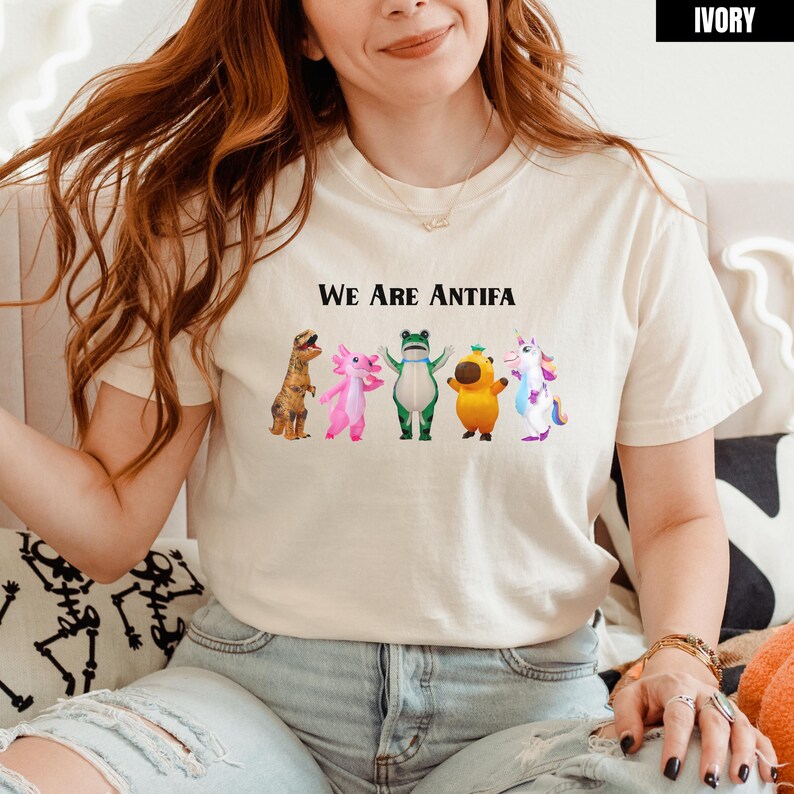 May include: Ivory-colored t-shirt with the text "We Are Antifa" above a row of cartoon characters: a dinosaur, axolotl, frog, a yellow creature, and a unicorn. The shirt is a crew neck and has short sleeves.