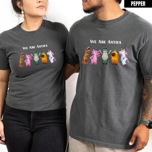 May include: Two charcoal gray t-shirts with the text "WE ARE ANTIFA" above a row of cartoon creatures. The creatures include a dinosaur, axolotl, frog, bird, and unicorn. The shirts are short-sleeved.