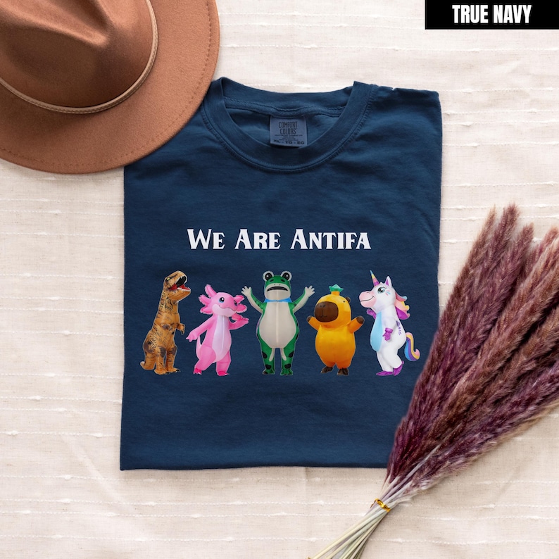 May include: True navy blue t-shirt with the text "We Are Antifa" above a row of cartoon animals. The animals include a dinosaur, axolotl, frog, chick, and unicorn. A brown hat and dried flowers are in the background.