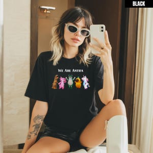 May include: Black t-shirt with the text "We Are Antifa" above a row of cartoon characters. The characters include a dinosaur, axolotl, frog, duck, and unicorn. The shirt is made of a soft material.