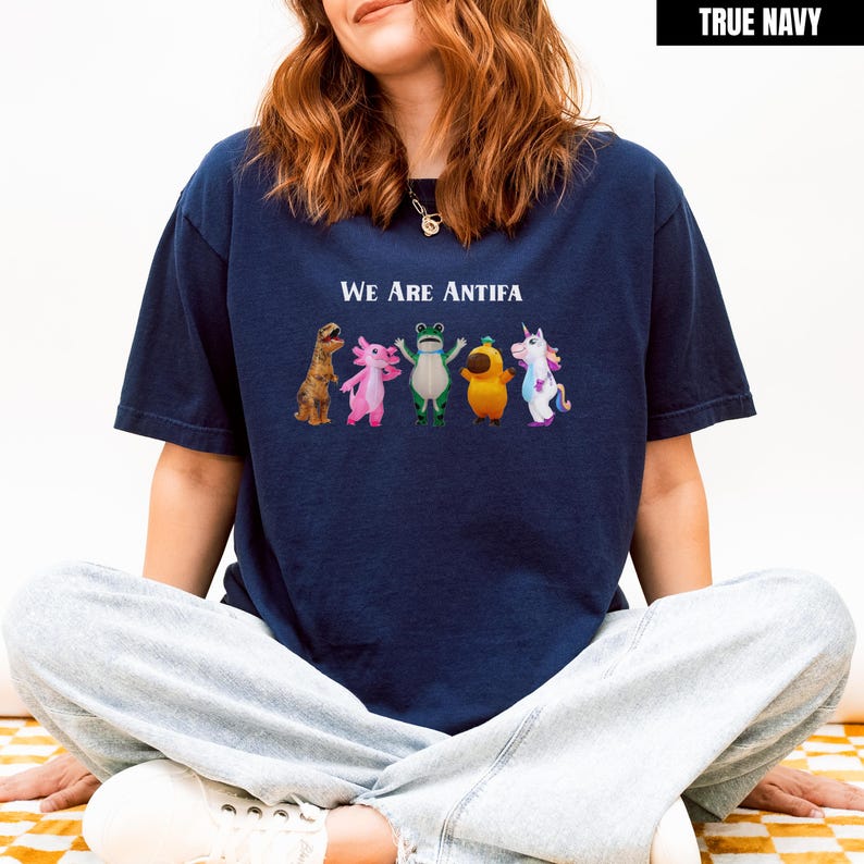 May include: Navy blue t-shirt with the text "WE ARE ANTIFA" above a row of cartoon characters including a dinosaur, axolotl, frog, bird, and unicorn. The shirt is a relaxed fit.