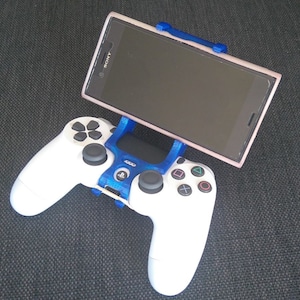 Może przedstawiać: Biały kontroler PlayStation 4 z niebieskim i czarnym uchwytem na telefon. Czarny smartfon ze srebrnym logo Sony znajduje się w uchwycie.