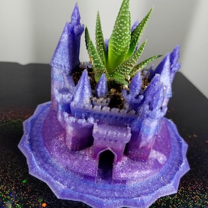 Puede incluir: Un macetero de castillo impreso en 3D de color morado y azul con una planta suculenta creciendo en su interior. El macetero está detallado con torres y una puerta.