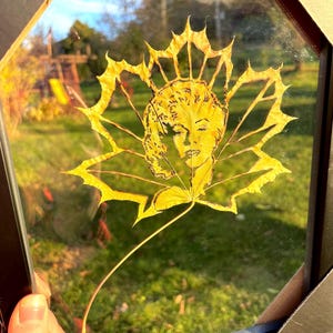 Op de afbeelding: Een geel blad met een gedetailleerde uitsnijding van een vrouwen gezicht, ingelijst in een zwart frame.