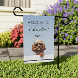 Può includere: Una bandiera da giardino blu e bianca con un cane marrone e bianco che indossa occhiali da sole. La bandiera recita "Benvenuti a casa di Charlie" con un'impronta di zampa nella parola "Home".