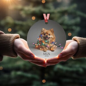 Round Christmas ornament.  Ornament has a Pomeranian wrapped in colorful Christmas lights.  Mia 2024 is at the bottom of the ornament.