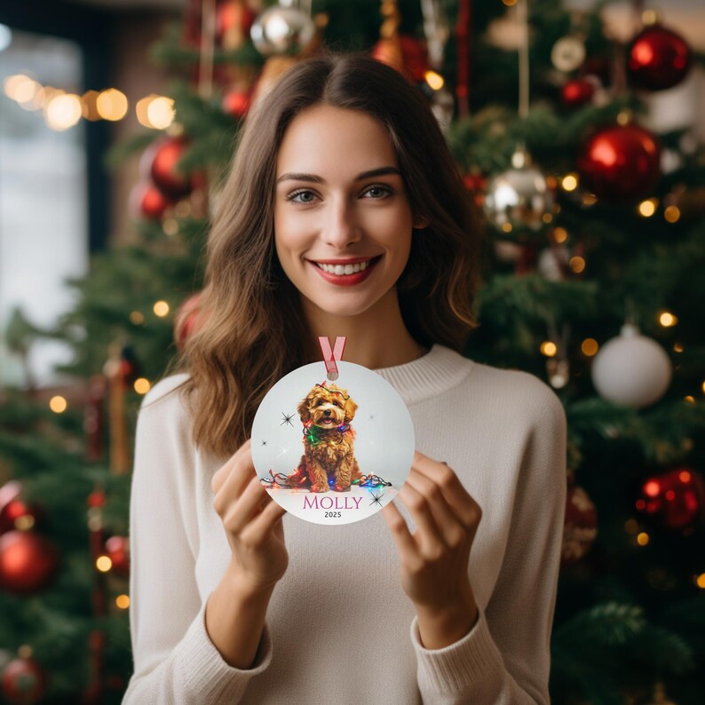 Woman is holding ornament in front of Christmas tree. Round aluminum Christmas ornament.  Ornament has a cute Cavapoo wrapped in colorful Christmas Lights.  Molly are in pink font and 2025 is in black font at the bottom of the ornament.  Names and year can be personalized.