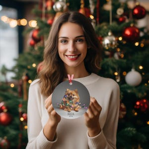 Round Christmas ornament.  Ornament has a Pomeranian wrapped in colorful Christmas lights.  Mia 2024 is at the bottom of the ornament.