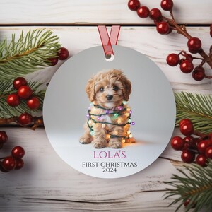 Op de afbeelding: Een ronde, witte kerstbal met een afbeelding van een kleine, lichtbruine puppy die in kleurrijke kerstverlichting is gewikkeld. De tekst "Lola's First Christmas 2024" staat onder de puppy.