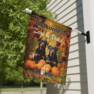 May include: Welcome to Zeus & Duke's Home garden flag with an autumnal theme. The flag features two black dogs, pumpkins, and autumn foliage.