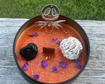 Könnte beinhalten: Ein schwarzes Kerzengefäß mit einer orangefarbenen Glitzerkerze darin. Die Kerze ist mit violetten Blumenakzenten, einer kleinen Schokoladentafel, einem schwarzen Stein und einem weißen Stein verziert. Ein silberner Jack Skellington-Ausschnitt sitzt oben auf der Kerze.