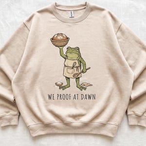 May include: A sand-colored crewneck sweatshirt featuring a graphic of a green frog wearing an apron, holding a basket of bread, and a wooden spoon. The text "WE PROOF AT DAWN" is printed below the frog, along with a small bowl and flour.