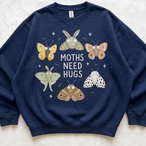 May include: A navy blue crewneck sweatshirt featuring a central graphic of five different illustrated moths surrounding the white text "MOTHS NEED HUGS" with small star accents. The long-sleeve pullover has a ribbed collar, cuffs, and hem.