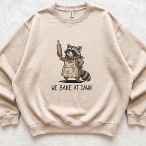 May include: A sand-colored crewneck sweatshirt featuring a graphic of a raccoon wearing a floral apron and a neckerchief, holding a wooden rolling pin. Below the illustration, the text reads WE BAKE AT DAWN in a simple, dark, sans-serif font.