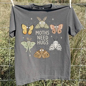 May include: A charcoal gray short-sleeve t-shirt featuring a graphic print of six cartoon moths in various colors and patterns surrounding the text "MOTHS NEED HUGS" in a white sans-serif font, with small white star accents scattered around the design.