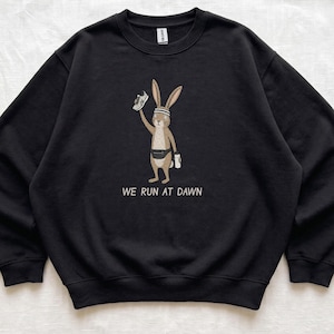 May include: A black crewneck sweatshirt featuring a graphic of a brown rabbit wearing a headband and a fanny pack, holding a running shoe and a water bottle, with the text WE RUN AT DAWN printed in white below the illustration.