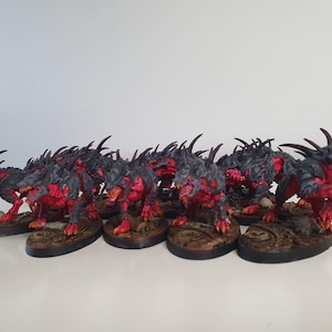 Puede incluir: Un grupo de 12 figuras en miniatura pintadas de negro y rojo con dientes y garras afiladas. Las figuras están en posición de carrera y están de pie sobre bases circulares.