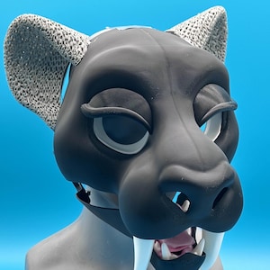 Puede incluir: Una máscara felina negra y gris con ojos grandes y prominentes colmillos blancos. La máscara presenta orejas grises texturizadas y está montada sobre una cabeza de maniquí gris sobre un fondo azul brillante.