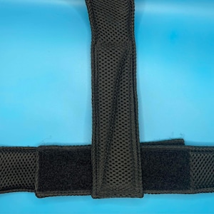 May include: A black mesh strap assembly featuring a vertical padded section connected to a horizontal band. The material has a textured, perforated surface with integrated hook-and-loop fastener panels for adjustable closure and secure attachment.