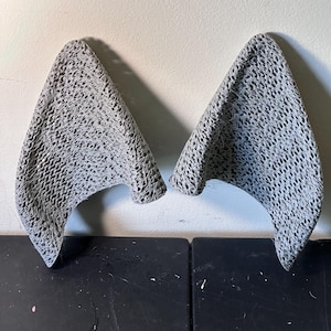 May include: A pair of gray fabric ears with a black and white patterned design. The ears are shaped like a cat's ears and are made of a soft, flexible material.