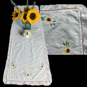 May include: White rectangular table runner with embroidered yellow and red floral designs. The runner has a crocheted lace border with a pink trim. A vase of sunflowers and a small white flower pot are on the runner.