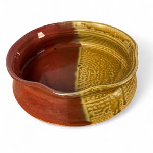 May include: A handmade ceramic bowl with a unique two-tone design. One half is a rich, reddish-brown, and the other is a warm, golden-yellow. The bowl has a slightly irregular shape, adding to its handcrafted charm. The interior features a textured pattern.