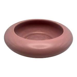 May include: A circular ceramic bowl with a smooth, matte finish in a dusty rose pink color. The bowl features a wide, rounded rim that curves inward toward a deep, concave center, creating a low-profile, minimalist vessel with a solid construction.