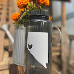 May include: A clear water bottle with a black lid and a white Nevada state outline with a heart cutout. The bottle is next to a small, gray watering can and a bouquet of bright orange flowers.